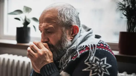 A man in a Christmas jumper huddles for warmth next to a heater
