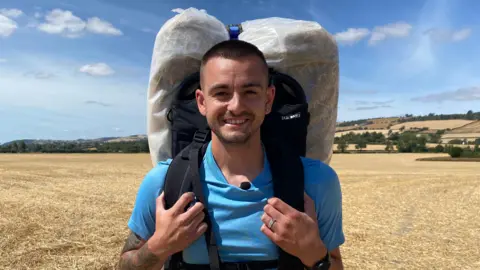 BBC Photograph of Firmstone, who is looking into the camera and smiling. He is wearing a bright blue T-shirt, and has a hay bale wrapped in plastic sheeting attached to a backpack on his back. He is holding onto the chest straps, and is stood on a yellow, stubbly field. The sky is bright blue and there is strong sunshine. 