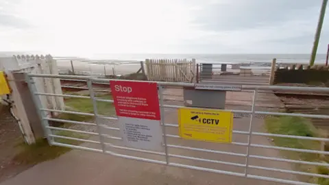 A general view of the level crossing at Braystones. There is a white gate with several signs warning to stop and that CCTV is in operation. The gate gives access to cross the line. On the other side is another gate leading to a road by the beach