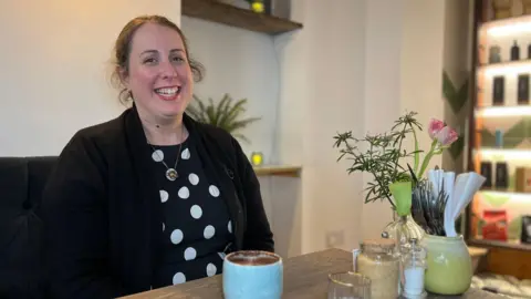 Sarah smiles at the camera as she sits at a table in a cafe. She has a black jacket on with a dark blue dress with white dots on under it. She has brown hair tied into a ponytail. There is a hot chocolate in front of her with flowers and cutlery also on the table.