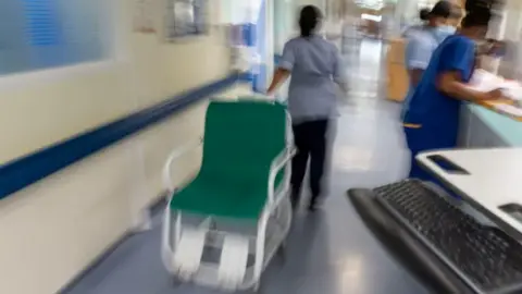 A nurse is moving a hospital chair along a corridor while two other nurses attend to paperwork