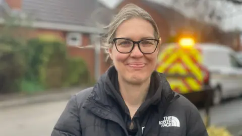 A woman with grey hair tied back who is wearing large black glasses and smiling into the camera. She is wearing a black North Face coat with a hoodie underneath. The background is blurred but you can see the side of a house and an emergency vehicle.