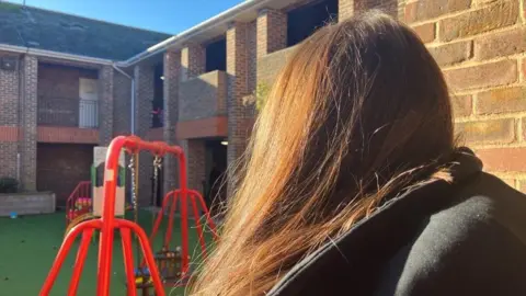 BBC Katrina has brown hair and a black coat. The background shows the playground which has red baby swings. 