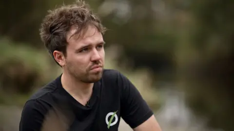 Matt Staniek, wearing a black T-shirt featuring the Save Windermere logo of an O resembling a sewage pipe with a green line slashed across it, pictured by the side of the lake.