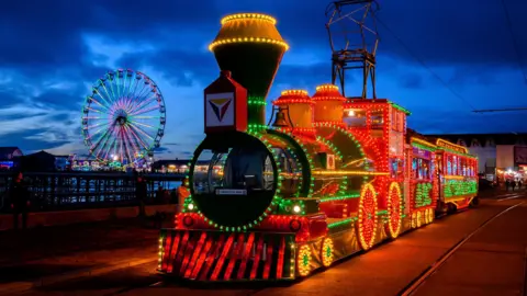 An old tram is covered in red, green and yellow lights travels along Blackpool Promenade at night. 