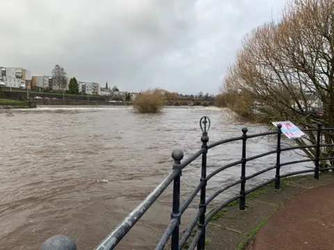 The River Nith in Dumfries with brown, murky waters and barriers around it and homes in the distance