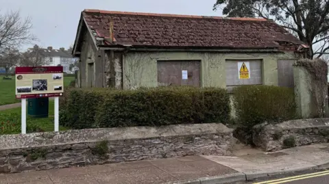 An old toilet block on Ropewalk Hill in Brixham, Devon. Its an old building painted green but has all its windows boarded up, with ivy and greenery growing up the side of it. Some of the bricks and tiles are falling offing the roof. 