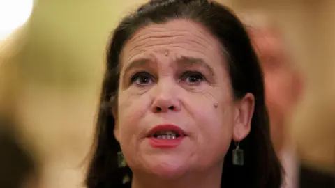 A woman with dark hair and earrings looks past the camera. In the background, there is another person, though they are out of focus. 