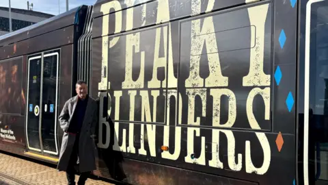 A man in a long grey coat stood next to a tram with the words Peaky Blinders emblazoned on the sides in cream lettering