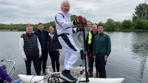 Tony Fahey, 82, who has white hair and is clean shaven, stands on a raft consisting of two white buoyant tubes and a metal frame. He is standing on the edge of a lake with a group of five men and one woman smiling behind him. 
