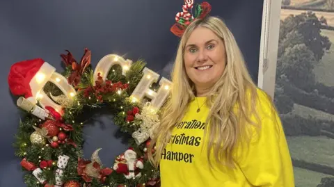 First West of England Charlotte has long, blonde hair and is wearing a festive headpiece and a yellow charity hoodie. She is standing in front of a Christmas wreath.