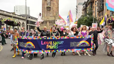 Organisers of Belfast Pride, and Lord Mayor Micky Murray, hold a banner with the organisation's banner on it with 'Love Your Mind' written in rainbow font. Many of the parade's participants are holding rainbow and progress flags, alongside the asexual and transgender flag.