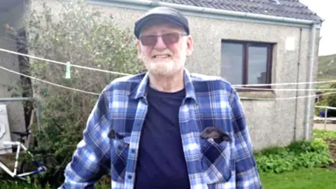 Margaret Mackay George Mackay standing in the back garden of a grey house. He is wearing a cap, a blue checked shirt, dark blue t-shirt and sunglasses, and has a white beard.
