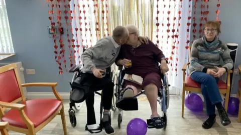 An elderly man and woman in wheelchairs share a kiss. The man is wearing a grey cardigan with a white shirt, the woman is wearing a purple/burgundy dress and a cross necklace. She is holding what appears to be a glass of orange juice while the man is also holding a glass of what appears to be cola. There are purple balloons on the floor, there is another woman to their left wearing a grey and blue striped jumper and navy blue jeans. There are red chairs with wooden frames and a blue wall in the background with love heart decorations that are hanging from the walls.