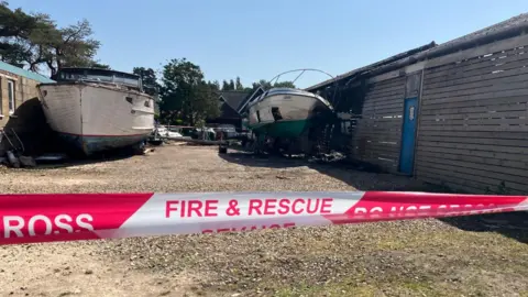 Owen Sennitt/BBC Tape cordons off a section of a boatyard that suffered a fire, with a badly damaged boat and boatshed behind.