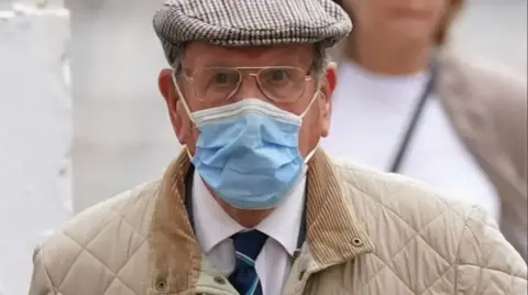 A man wearing a white shirt, navy tie and cream coat. He is also wearing a blue facemask.
