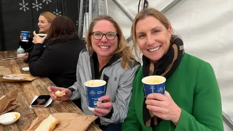 BBC Liz Casely (Left) and Flo Highland (right) were among the happy customers at the annual event. Liz wears a reflective, silver waterproof jacket, while Flo is wearing a green coat and a black and beige scarf. Both have golden hair and are holding their paper cups of soup and smiling