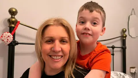 A woman smiles as she sits on a bed with her son, who has his arms around her. The woman has strawberry-blond hair, a silver nose stud and wears a black T-shirt. The boy has short blond hair and wears a red T-shirt. The bed has a black and gold frame. The wall is cream-coloured.