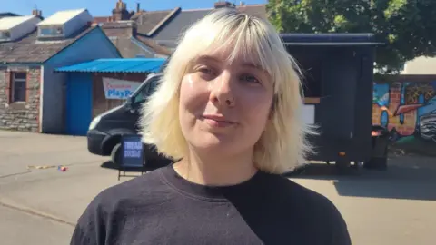 Izzy Cross is seen from her shoulders up standing in front of a black van. She has short, blonde hair and is smiling.