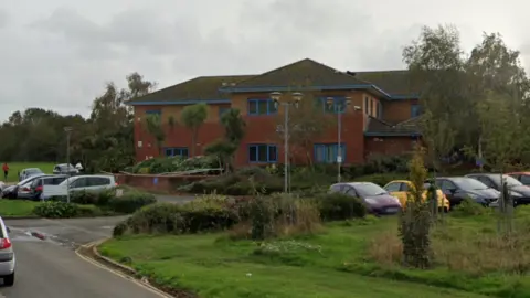 A red brick building with blue windows is seen from a short distance. The entrance to the car park, which is full of cars, isalso  visible. 