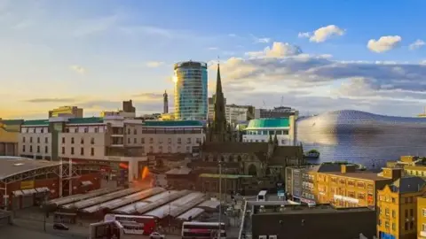 A general view of Birmingham. At the front are buses and behind them market stalls. A church with a large steeple rises up and behind that a circular tall building. The sky is sunny and it looks like sunset.
