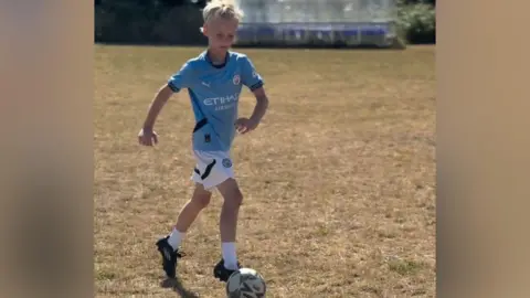 Handout A blond boy wearing a blue football jersey and white shorts, is dribbling a football on a football pitch. 
