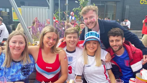 Sam O'Brien West Sam, with England flags painted on his cheeks, smiling into the camera as he poses with five of his friends, all wearing England football T-shirts, hats and face paint. They're sitting outside in a beer garden, with several alcoholic drinks on the able in front of them.