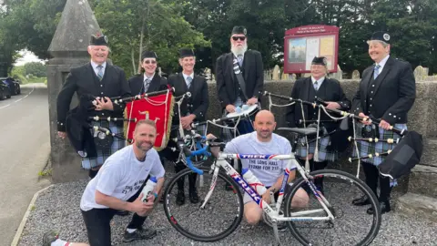 JODIE CANNELL Darren and Adrian wear white T-shirts and pose with their bikes in front of Ellan Vannin Pipes and Drums who wear kilts, one has a Manx flag, and they have drums and bagpipes.