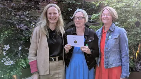 Jen Donnelly Three women are smiling. A woman in the middle with glasses is holding a paper envelope with the medal encased.