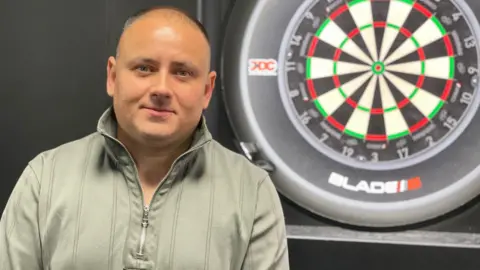 Steve Brown looking at the camera. He has short dark hair and is wearing a long-sleeved zip-up top. He is standing in front of a dart board.
