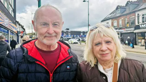 A man and woman stand together looking at the camera. He has short grey hair and a moustache, and wears a black padded coat with a red lining and a maroom jumper underneath. His wife has blonde shoulder length hair and sunglasses on her head. She has a brown padded coat on, a cream jumper underneath and a necklace. Behind them is a row of shops and a main road.