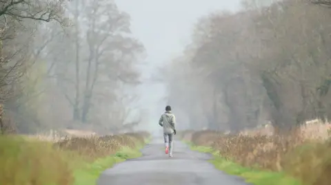 PA Media A person out jogging on a foggy morning. They are pictured with their back to the camera. They are wearing a black beanie, a beige jacket, black gloves, grey trousers and trainers with bright orange soles. They're travelling down a lane in Glastonbury which is lined by trees and grass.