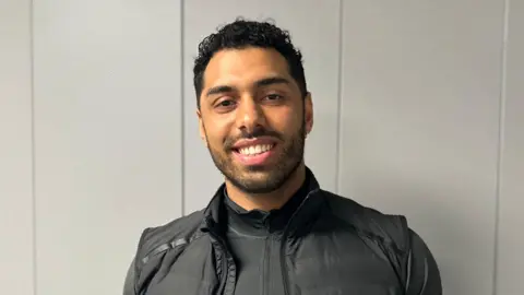 A picture of Leon Greenwood in the BBC Radio Berkshire offices. He is wearing a black zip-up top with a black gilet. He has short dark hair and is looking at the camera and smiling.