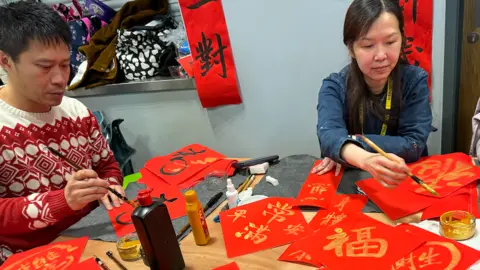 Three people write gold calligraphy onto red banners for Lunar New Year