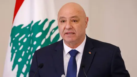 Getty Images Joseph Aoun, a bald man wearing a blue suit and blue tie with white shirt stands in front of the Lebanese flag.