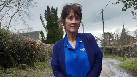 LDRS A woman wearing a nursing uniform and a cardigan, with brown hair and a fringe and wearing glasses on her head, stands on a lane with hedges on either side and houses and a church behind her.