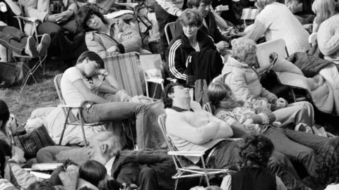 Getty Images A black-and-white image of people in rows of deckchairs, some sleeping, with bags and coats strewn around them.