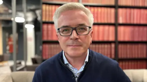 Alun is wearing dark-framed glasses and has a neutral expression. He is wearing a light blue checked shirt with a navy sweater. Behind him, blurred, are red books on a large bookshelf. It is a head and shoulders shot of him.