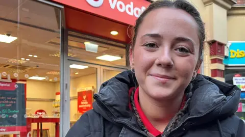 Grace Pearson with brown eyes and hair in two French plaits wearing a black coat and red polo top. She is sitting on a bench outside a mobile phone shop. She is smiling.