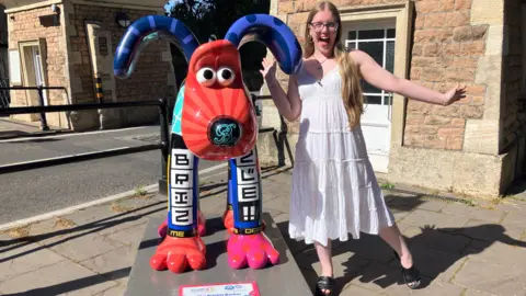 Naomi, aged 17, stands smiling beside a brightly coloured Gromit sculpture. She is wearing a white dress and striking the same playful pose she did as a young child. The sculpture features vivid patterns and cheerful colours.