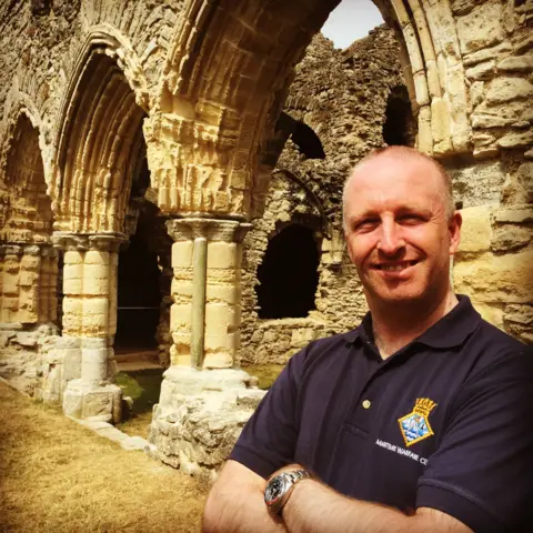 Richard Jones Richard Jones standing in front of historic ruins. He has his arms folded and is wearing a black polo shirt with a logo on it. He is bald and is smiling at the camera.