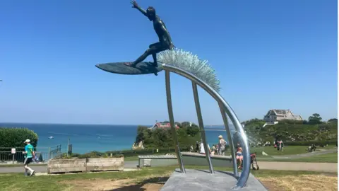 A bronze statue of a surfer wave riding. It's dark and taken on a bright sunny day with blue skies in Newquay. The sea is visible in the background along with some homes and grass. 