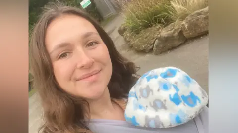 Megan Meiring smiles at the camera in a selfie-style image. She has long brown hair and holds a baby on her chest wrapped in sling while it has a blue and white hat over its head.