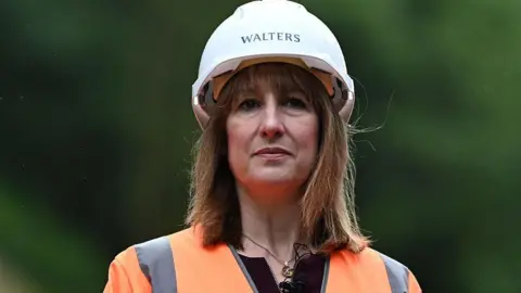 Rachel Reeves, who has brown shoulder-length hair, is wearing an orange high-vis jacket and a white hard hat