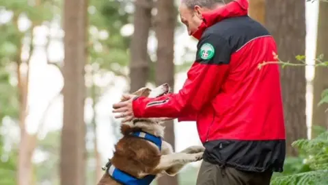 Surrey Search & Rescue A man in a red jacket bending down and speaking to a dog