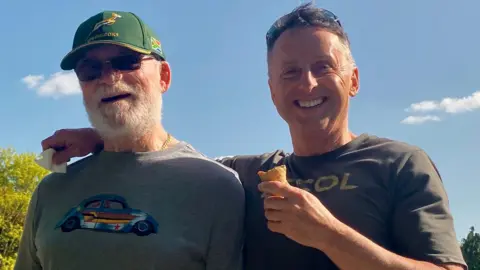 Andy North Two men wearing grey t-shirts, one wearing a cap and one eating an ice cream, against a blue sky.