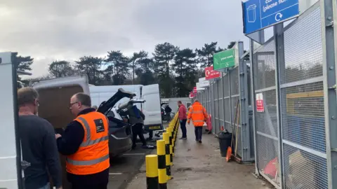 Mousumi Bakshi/BBC Vehicles are pictured parked up at the entrance of recycling centre's bins. Various areas for different items are marked out by signs. Centre workers in hi-vis jackets can be seen moving around and talking with customers.