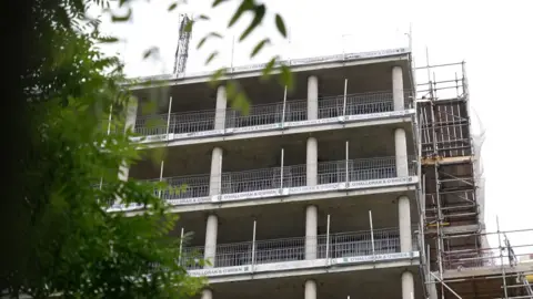 A housing development under construction. Concrete storeys are installed with wire safety netting at the side. Scaffolding goes up the right side of the building.