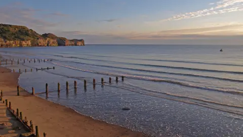 Geograph/WF Millar Sandsend beach at sunset, with groynes reaching out into the low tide and a boat in the distance.