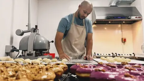 SAY Doughnuts Aaron Shade, making doughnuts, in a purpose built kitchen, he is by a food mixer, and an oven behind him. with row is doughnuts in front of him, with different toppings. He has on a beige apron, blue T-shirt. He is looking down.